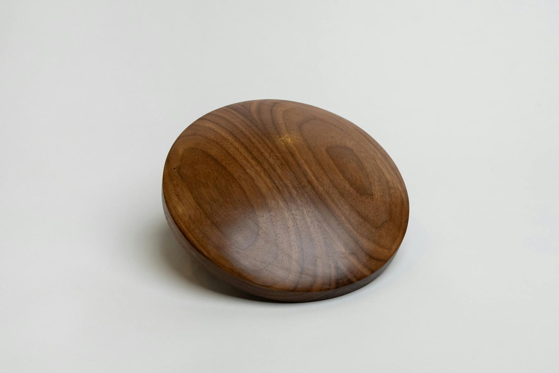 A thin, walnut saucer with both the top and bottom bulging outward. Its surface is smooth, showing faint reflections of the studio lights around it.